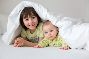 Cute little six month old baby boy and his older brother, playing under duvet at home in bed in bedroom, smiling