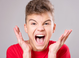 Emotional portrait of irritated shouting teen boy. Furious teenager screaming and looking with anger at camera. Handsome outraged child shouting out loud on gray background.