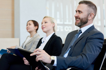 Group of smiling business people sitting in row in audience listening to training seminar, focus on handsome young businessman in front