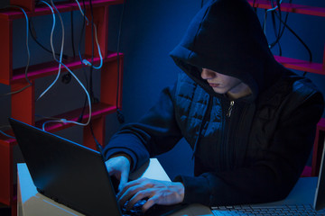 A hacker in a hood sits in a dark room with a black laptop
