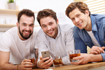 Cheerful friends having fun with smartphone and drinking at home