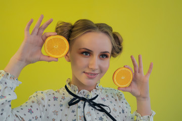 Portrait of a blonde girl with a funny hairdo and in a light shirt holding slices of oranges posing against a bright background