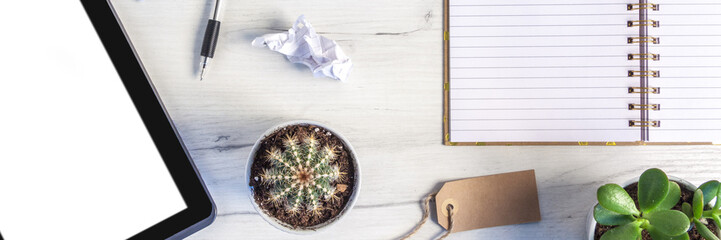 Panoramic top view on a white desk with an open notebook, tablet, plants and crumpled sheets of paper