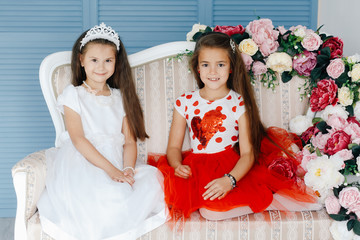 Two little beautiful princesses in luxurious dresses posing on sofa with flowers nearby