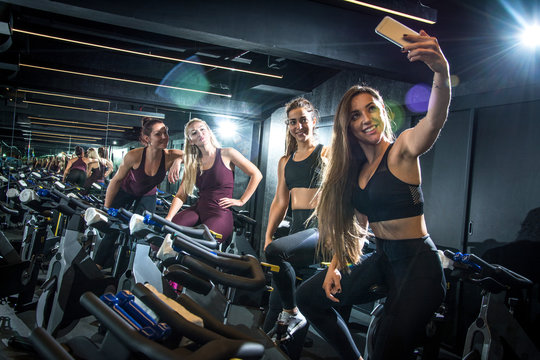 Sporty girls taking selfie while sitting on exercise bikes in gym