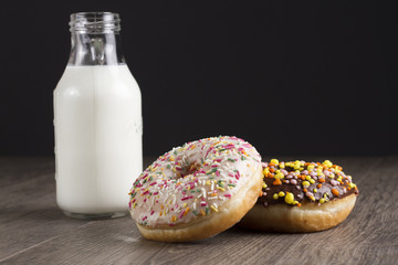 Two donuts lying on the table and on a black background. A bottle of milk is next to donuts on a wooden table. Space for text.