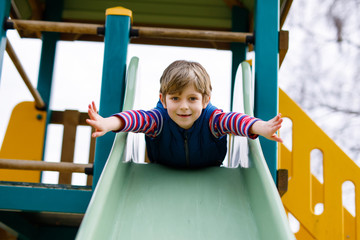 Happy blond kid boy having fun and sliding on outdoor playground