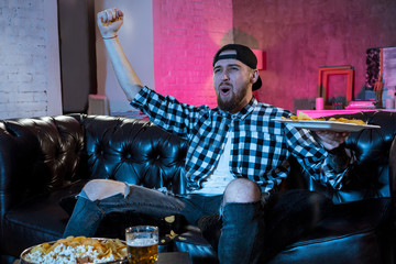 A handsome bearded man watching a football match on TV sitting at home on the couch and drinking beer.