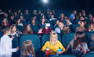 Cinema hall full of young people and children,looking surprised and exited, watching on girl wearing 3D-eyeglasses. People sitting on comfortable plases feeling interested.