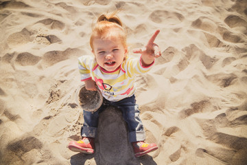 Portrait of small baby, little girl in blue jeans, pink shoes and colourful pullover sitting and playing in sand at the beach, top view