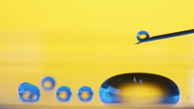 A hilarious macro of wee drops of liquid dribling from a metallic needle and sliding on big round pool in the yellow background of a lab.