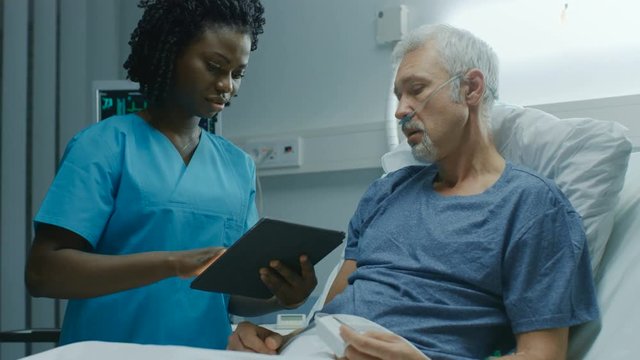 In the Hospital, Senior Patient Lying in the Bed Talking to a Nurse who is Holding Tablet Computer Showing Him Information. Shot on RED EPIC-W 8K Helium Cinema Camera.
