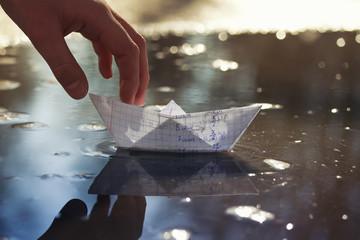 childrens hand of the schoolboy launches a small paper boat made of a school notebook in a spring early puddle on a Sunny bright day