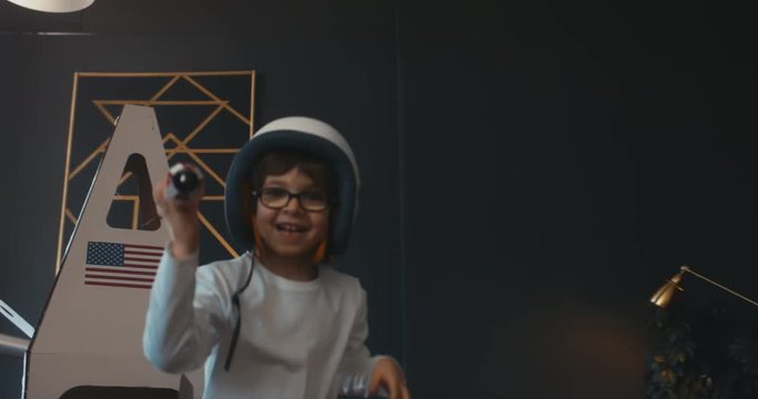 TRACKING Cute little kid boy wearing astronaut helmet getting out cardboard spaceship and holding toy rocket in his hands. 4K UHD 60 FPS SLOW MO