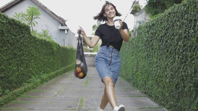 Young woman holds string shopping bag and reusable cup with hot drink. Happy girl run at suburban street to home. Zero waste noplastic concept. Slow motion video shooting by handheld gimbal