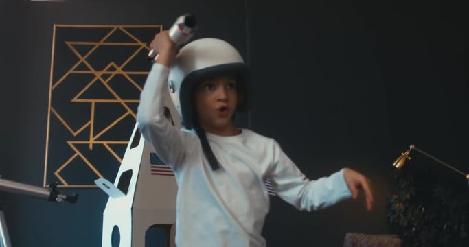 TRACKING Cute little kid boy wearing astronaut helmet getting out cardboard spaceship and holding toy rocket in his hands. 4K UHD 60 FPS SLOW MO
