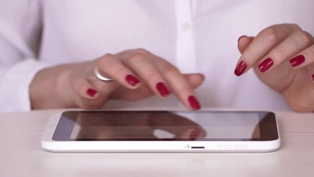 The woman in a white shirt watches information on the tablet. The businesswoman works at office or at home. Typing on the tablet. Hands close up. hand touching tablet computer surface touchscreen