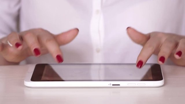 The woman in a white shirt watches information on the tablet. The businesswoman works at office or at home. Typing on the tablet. Hands close up. Beautiful red manicure close up. hand touching tablet