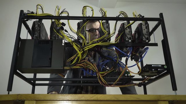 Young male person in work coat maintains a mining rig machine with six graphics cards obviously not satisfied with the condition what state they are, concept cryptocurrency computer, symbolic shot.