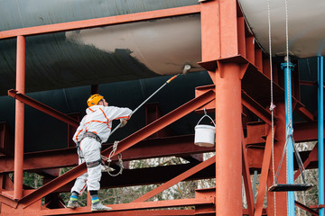 Naklejka premium Industrial climber in helmet and uniform painting water tower. Professional Painter working on height. Risky job. Extreme occupation.