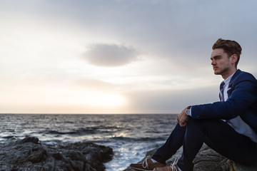 Serious young man in elegant suit sits on the coast near ocean. Young guy thinking on background of the water and sky