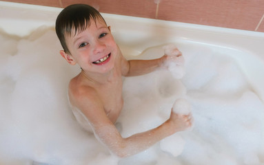 Boy 6-7 years sitting in the bath with foam looking into the camera and smiles. View top.