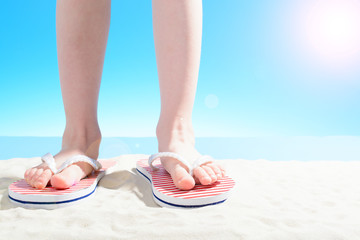 A little girl in flip flops stands on a sandy beach. body part