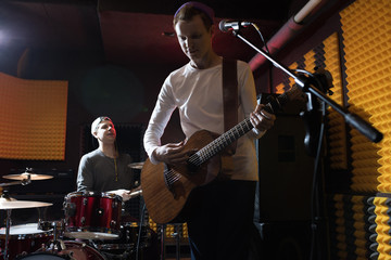 Band of young musicians performing in dim recording studio making new album, singer with guitar in foreground, copy space