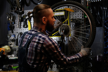 Fototapeta na wymiar Handsome stylish male wearing a flannel shirt and jeans coverall, working with a bicycle wheel in a repair shop.