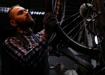 Naklejka na ściany i meble Handsome stylish male wearing a flannel shirt and jeans coverall, working with a bicycle wheel in a repair shop.