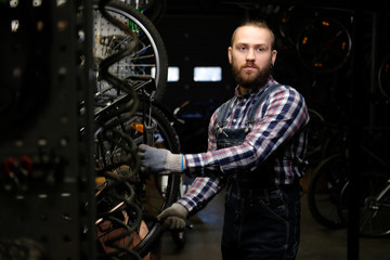 Handsome stylish male wearing a flannel shirt and jeans coverall, working with a bicycle wheel in a repair shop. A worker using a wrench mounts the wheel on a bike in a workshop.
