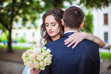 Bride hugs groom by shoulders, holding in her hand wedding bouquet of roses