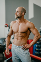 Fototapeta na wymiar Shredded shirtless tough bodybuilder with beard watching to the left side, posing in a gym.