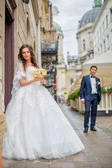 Naklejka na ściany i meble Beautiful bride in white wedding dress is holding bouquet of roses, behind her is groom in tuxedo. Romantic walk around the city