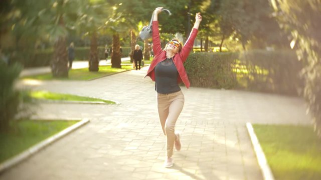 a woman in sunglasses and a red jacket dancing and jumping over the city park on a clear sunny day. 4k, slow motion