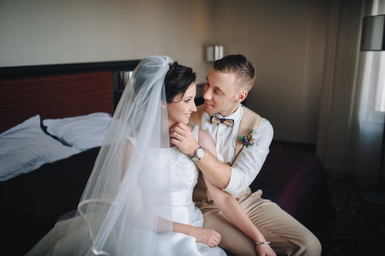 Beautiful newlyweds are sitting in a chic hotel room. The bride and groom embrace in the room, sitting on the bed.