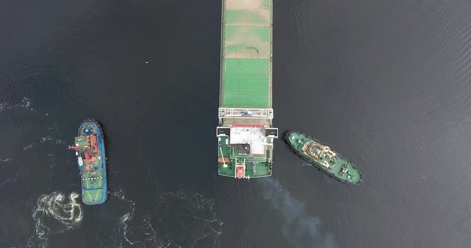 River tug helps when mooring a dry cargo tanker on a calm river early in the morning. Summer day. The camera moves in the air near from the ship. Aerial view.