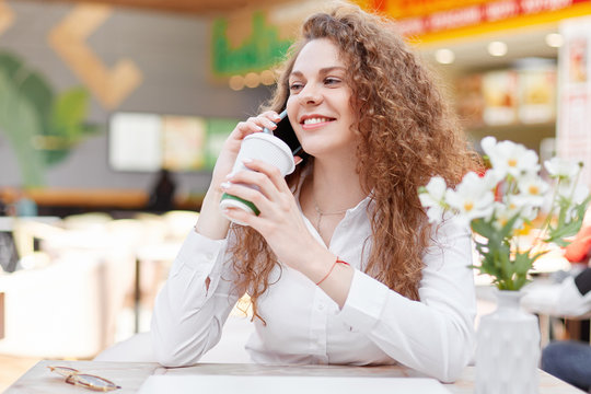 Lovely good looking curly female model speaks via cell phone with boyfriend, shares positive news, has coffee break after walk in nearest cafe, looks joyfully into distance. People, communication