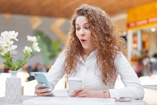 Portrait of amazed woman looks with surprised expression in smart phone, doesn`t expect to recieve message from male, sits at table in cafe, decorated with flowers, drinks coffee. Wow, how wonderful