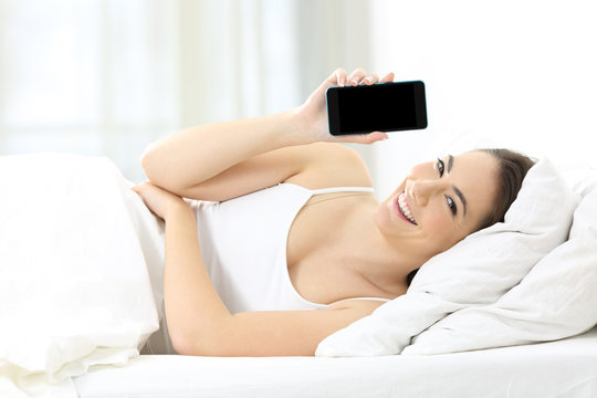Happy woman showing a blank phone screen in the bed