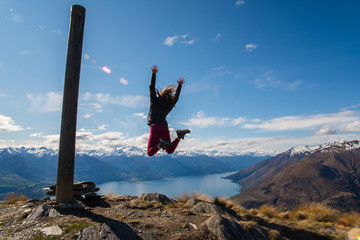 A woman jumping up in the air on the summit of a mountain with a lake in the background. 