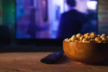 Naklejka na ściany i meble A wooden bowl of popcorn and remote control in the background the TV works. Evening cozy watching a movie or TV series at home