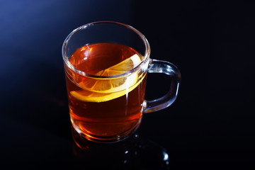 A glass mug of tea with lemon stands on a black glass. A cup of tea is beautifully reflected in the glass