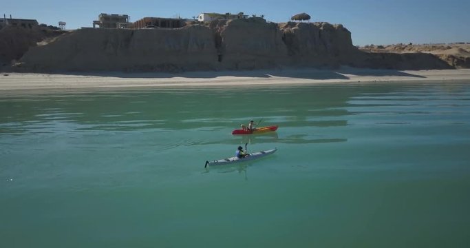 Two female kayakers paddling along a beach with steep sandy cliffs.