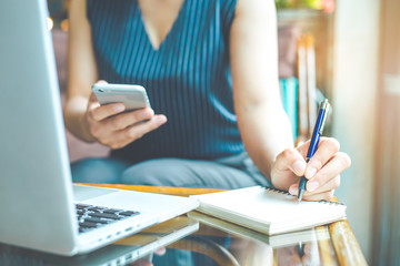 Business woman hand is writing on a notepad with a pen and using a mobile phone.