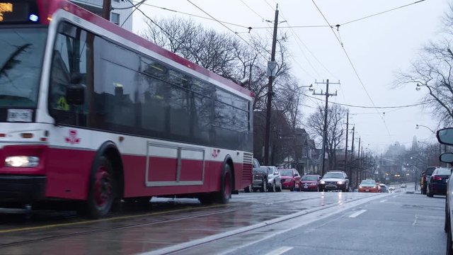 I Toronto city bus passes by on a foggy rainy day. We are on a residential street and we can see streetcar wires overhead.
