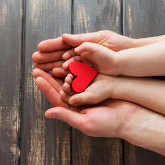 Two pairs of hands, men and children on a wooden background. Four hands are holding a red, bright heart.