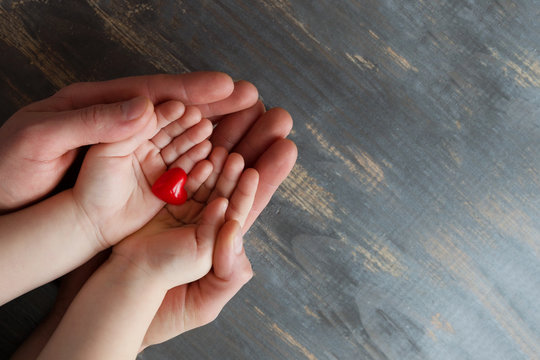 Two pairs of hands, men and children on a wooden background. They hold a tiny red heart in their hands.