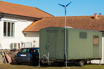 Repurposed military trailer outside a house. Small wind power generator supply electricity. Car parked beside trailer. Logo and registration removed.