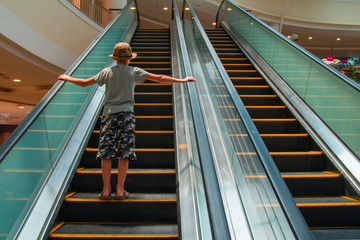 Boy rises up on an escalator in shopping center. Child in hat, t-shirt and breeches. Rear view. Copy space.